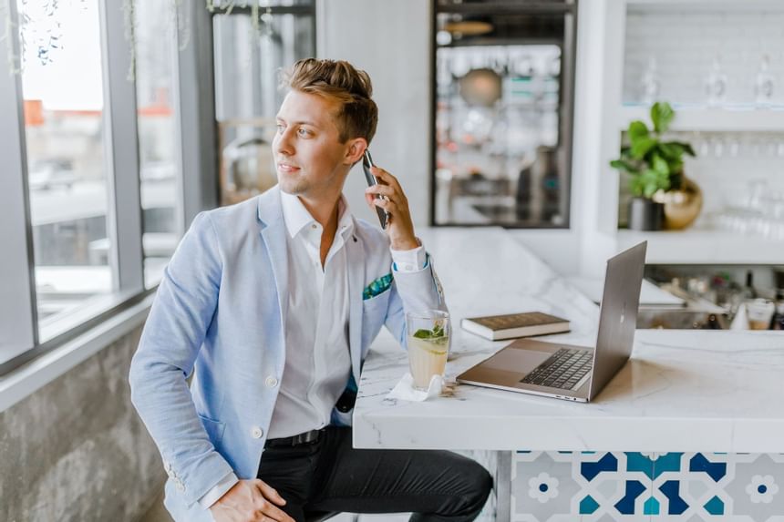 Man using a laptop beside a drink on a marble counter by bright windows in a modern workspace at Hotel Saint Clair