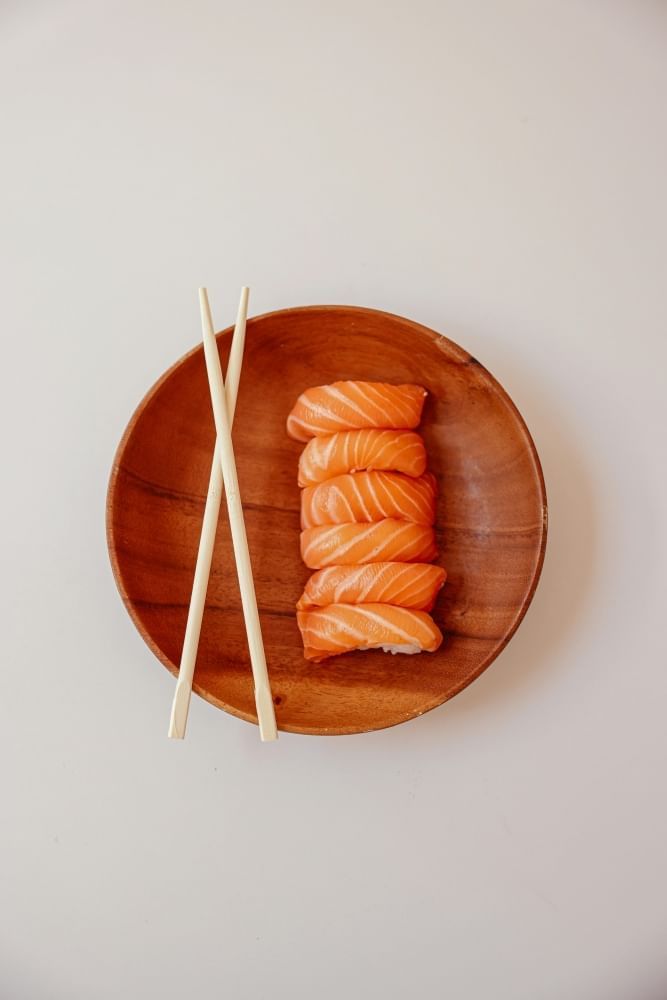 A simple wooden bowl with crossed chopsticks and six slices of salmon nigiri against a gray backdrop.  