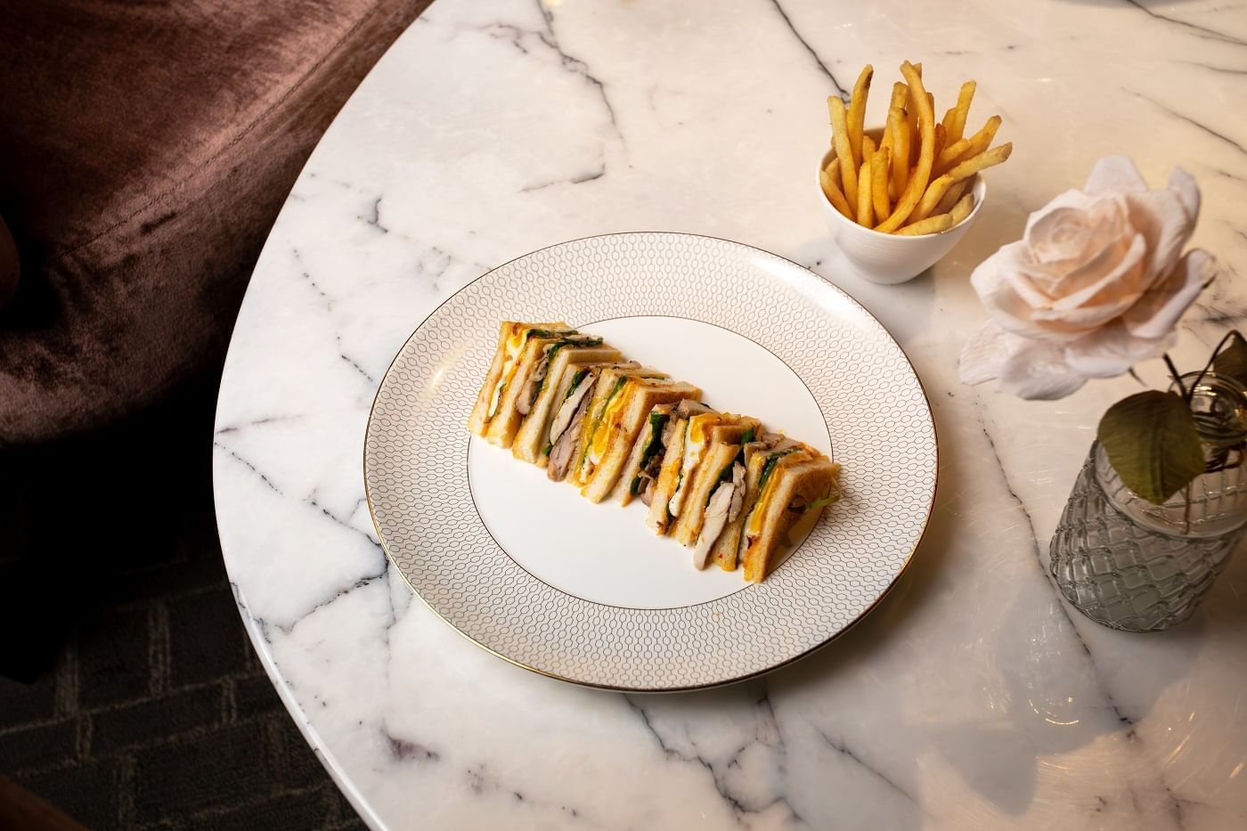 A perfectly cut club sandwich with golden fries and a rose on a marble table at The Londoner Hotel