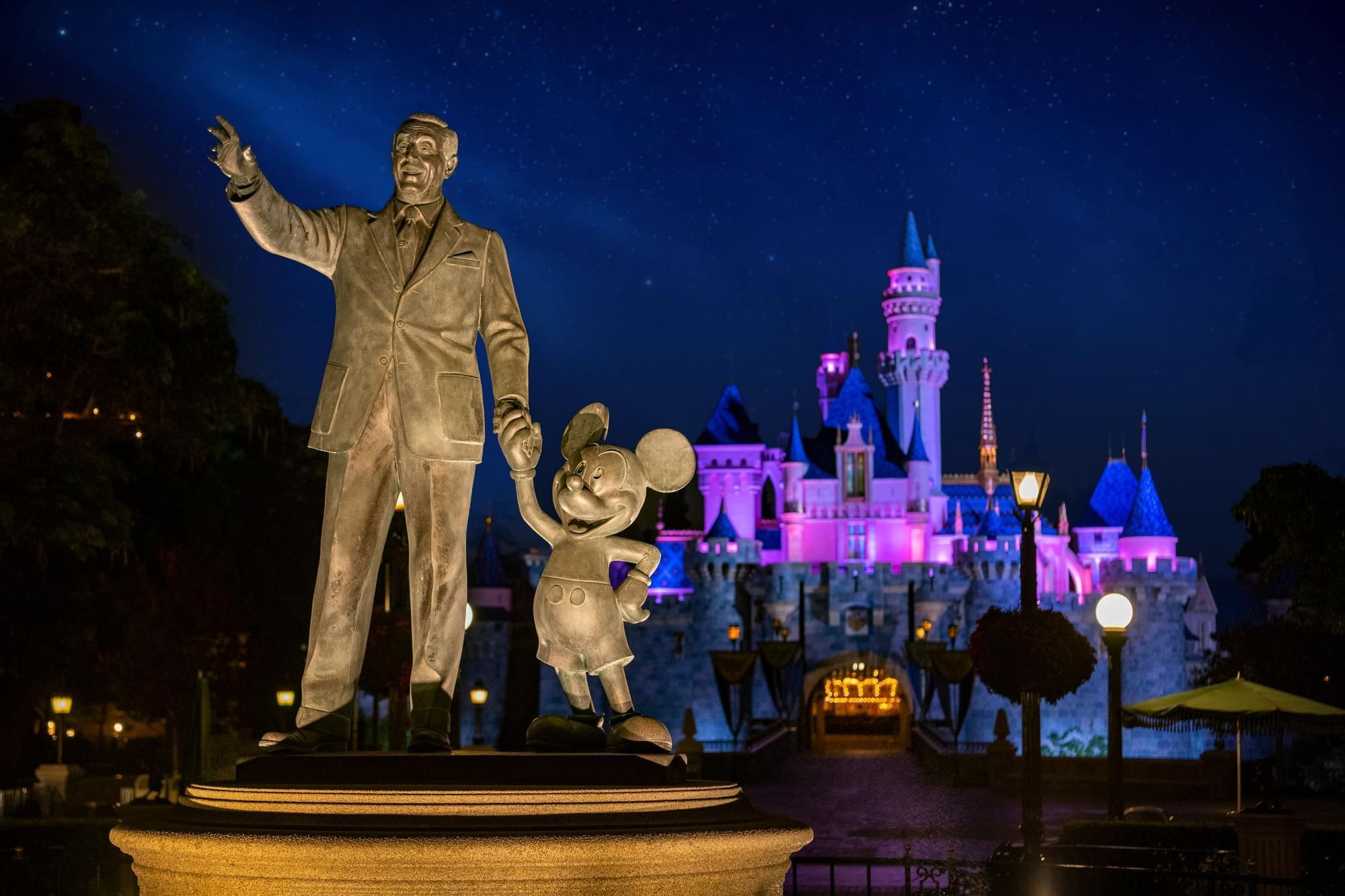 A statue of Walt Disney and Mickey Mouse in front of a castle at night.