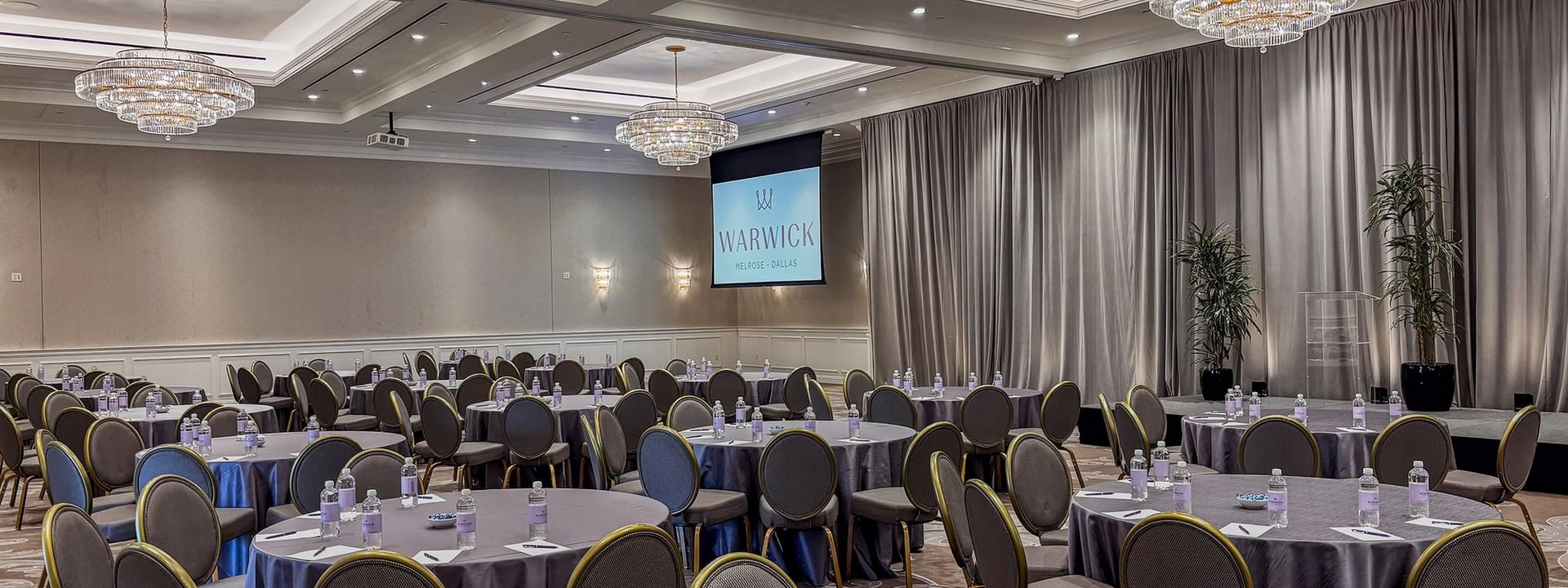 Round tables by grey chairs under crystal chandeliers in Turtle Creek Ballroom at Warwick Melrose Dallas