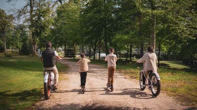 A family of four rides bicycles on a dirt path through a park, surrounded by trees.
