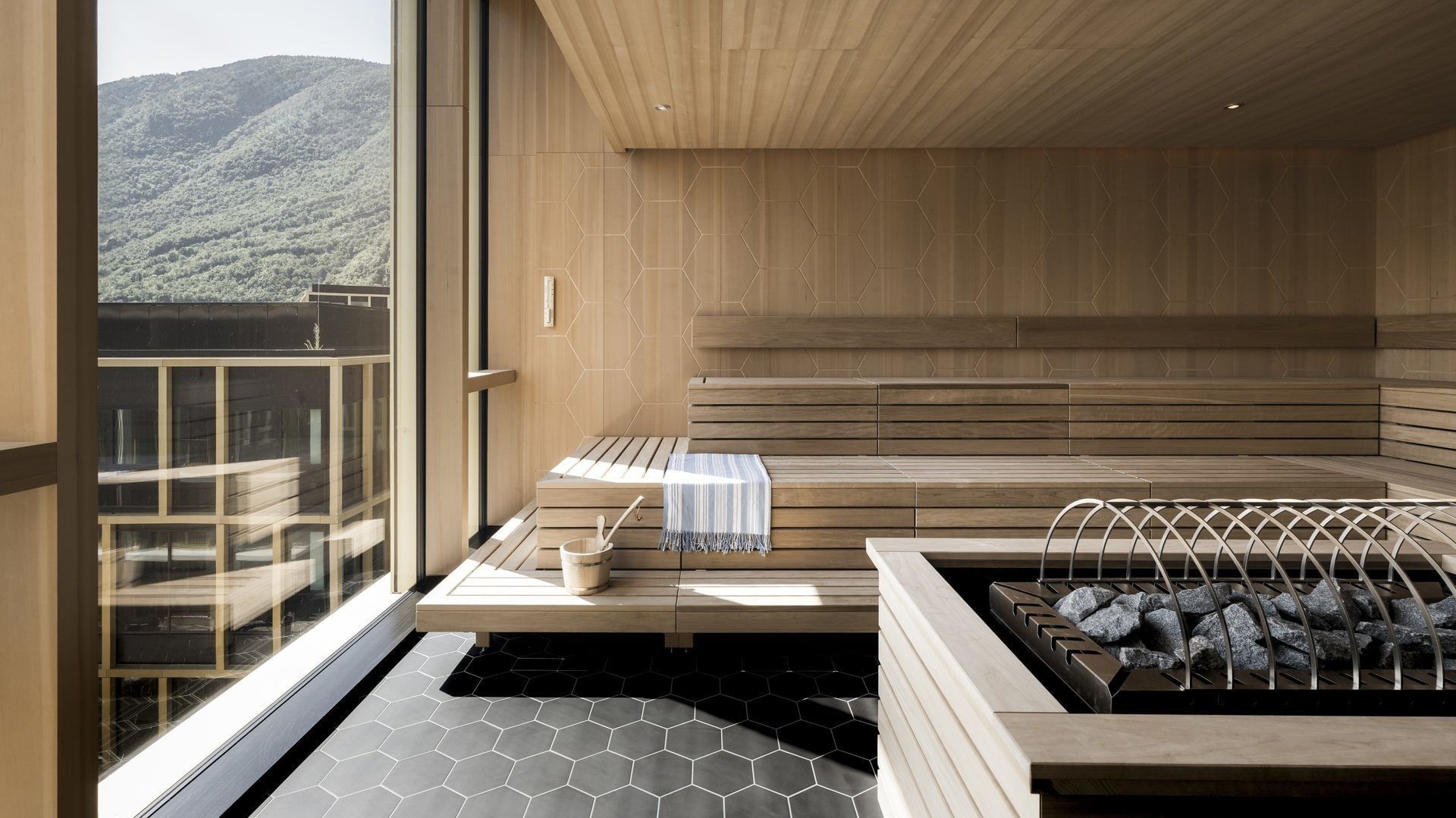Sauna in legno con vista su montagne e struttura moderna, pavimento esagonale nero.