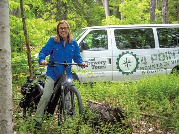 Woman on mountain bike beside 4 Points VT tour van in lush green forest setting