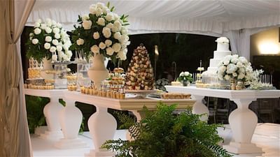 A dessert table with white floral arrangements, including a large white wedding cake and small pastries at Hotel Mykonos