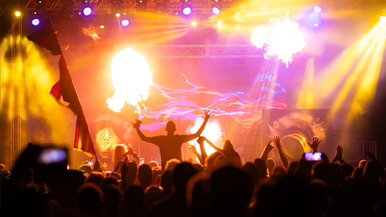 Performer on stage with raised arms, surrounded by flames and colorful lights, crowd below.