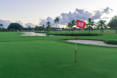 Red 18 flag on a putting green near a sand bunker and pond with palm trees in Miami Lakes Golf Club at Miami Lakes Hotel