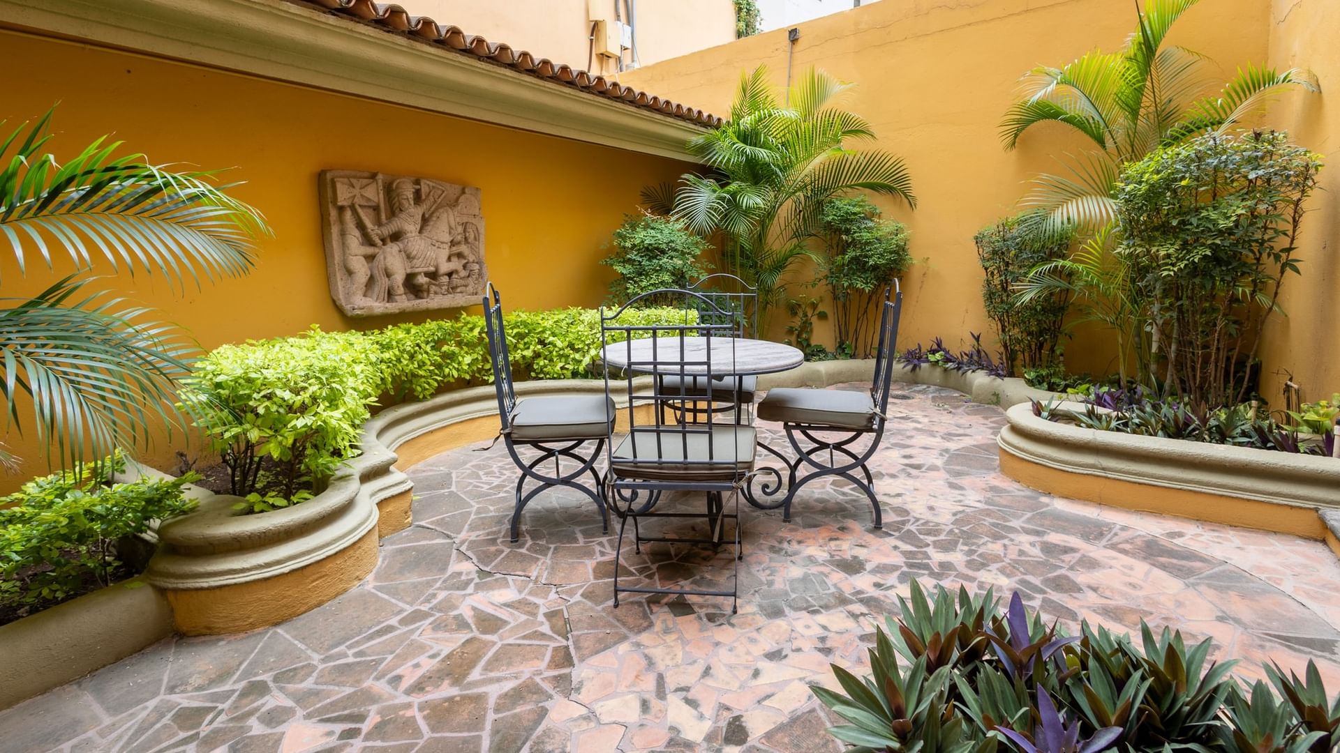 Patio privado con mesa y sillas de hierro forjado, jardineras con vegetación tropical y paredes amarillas en Quinta Real Monterrey.