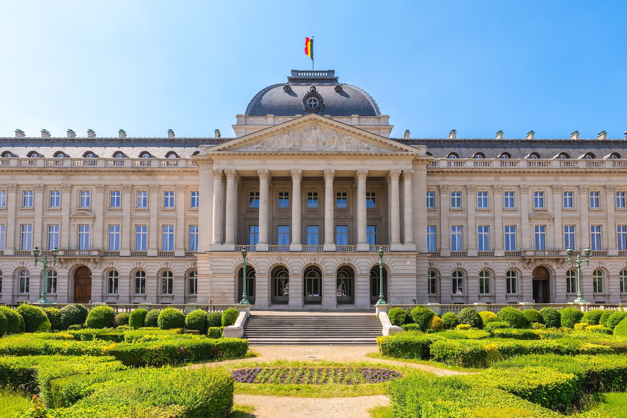 Grand facade of the Royal Palace, with flags flying, surrounded by manicured gardens near Warwick Grand Place Brussels