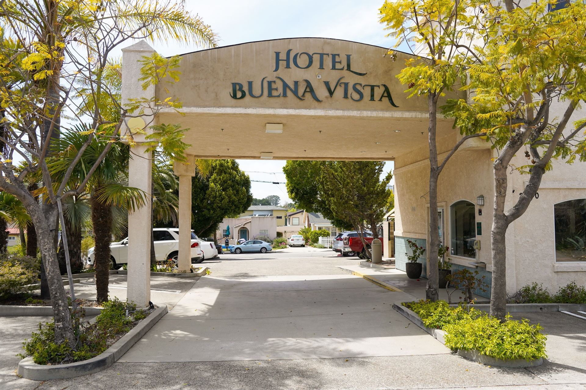 A beige archway with 'Hotel Buena Vista' in black letters stands at the entrance of a hotel, with trees and cars visible.