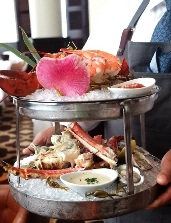 A waiter presents a two-tiered seafood platter, adorned with lobster, shrimp, crab legs, and oysters at The Diplomat Resort