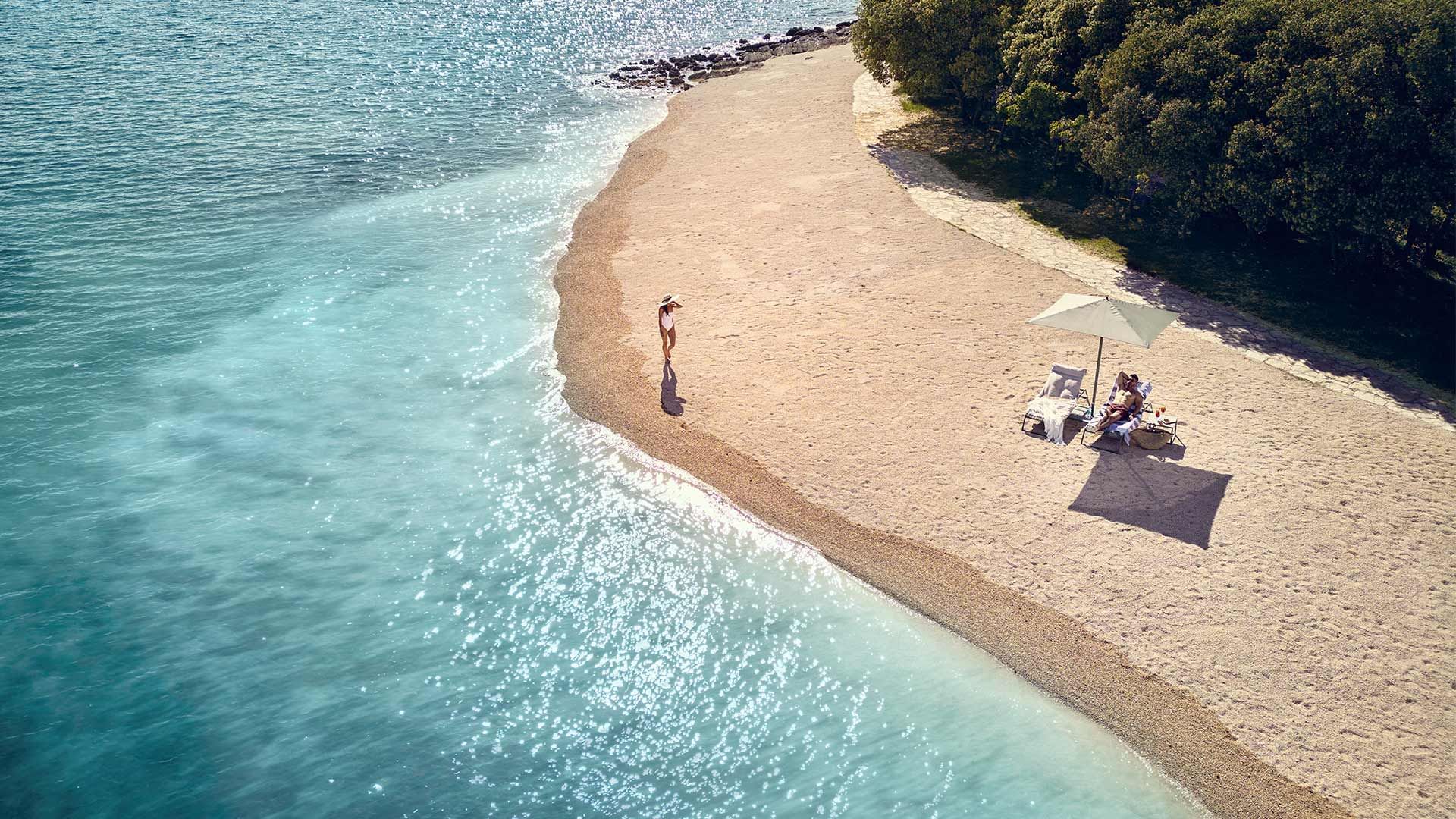 Iz zraka, osoba hoda plažom uz more s ležaljkama i suncobranom.
