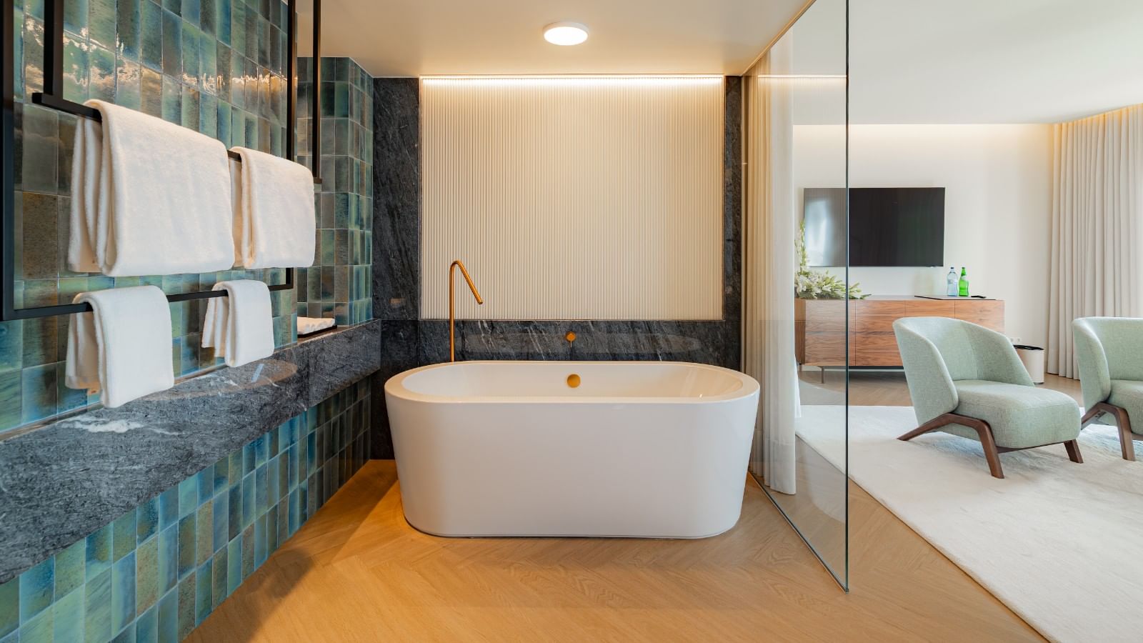 Banheiro com banheira, toalhas, paredes de azulejos azuis e verdes, e cadeiras verdes no quarto adjacente.