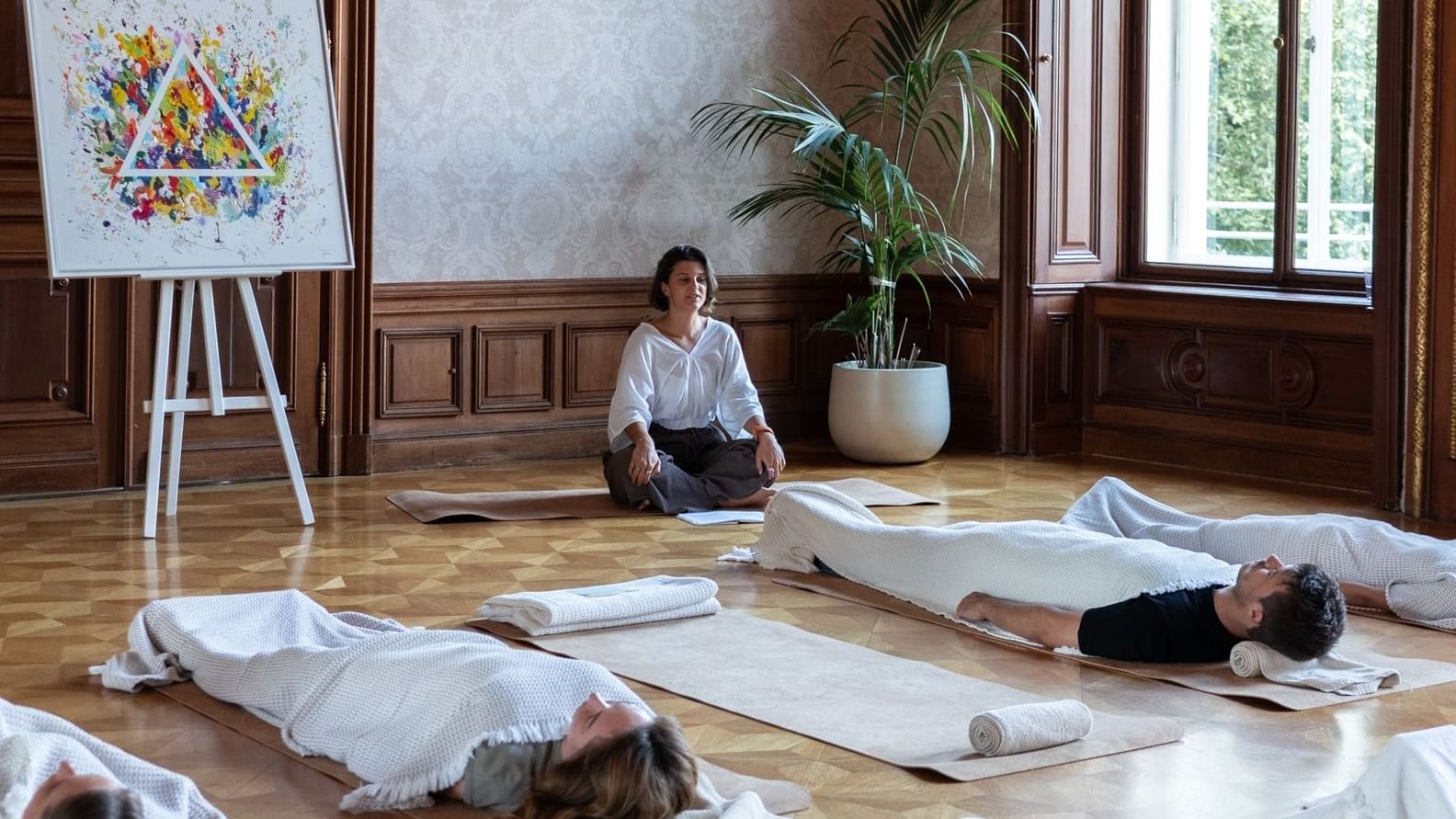 Participants lying on mats with blankets in a room with a colorful painting and lush plants at Almanac Palais Vienna