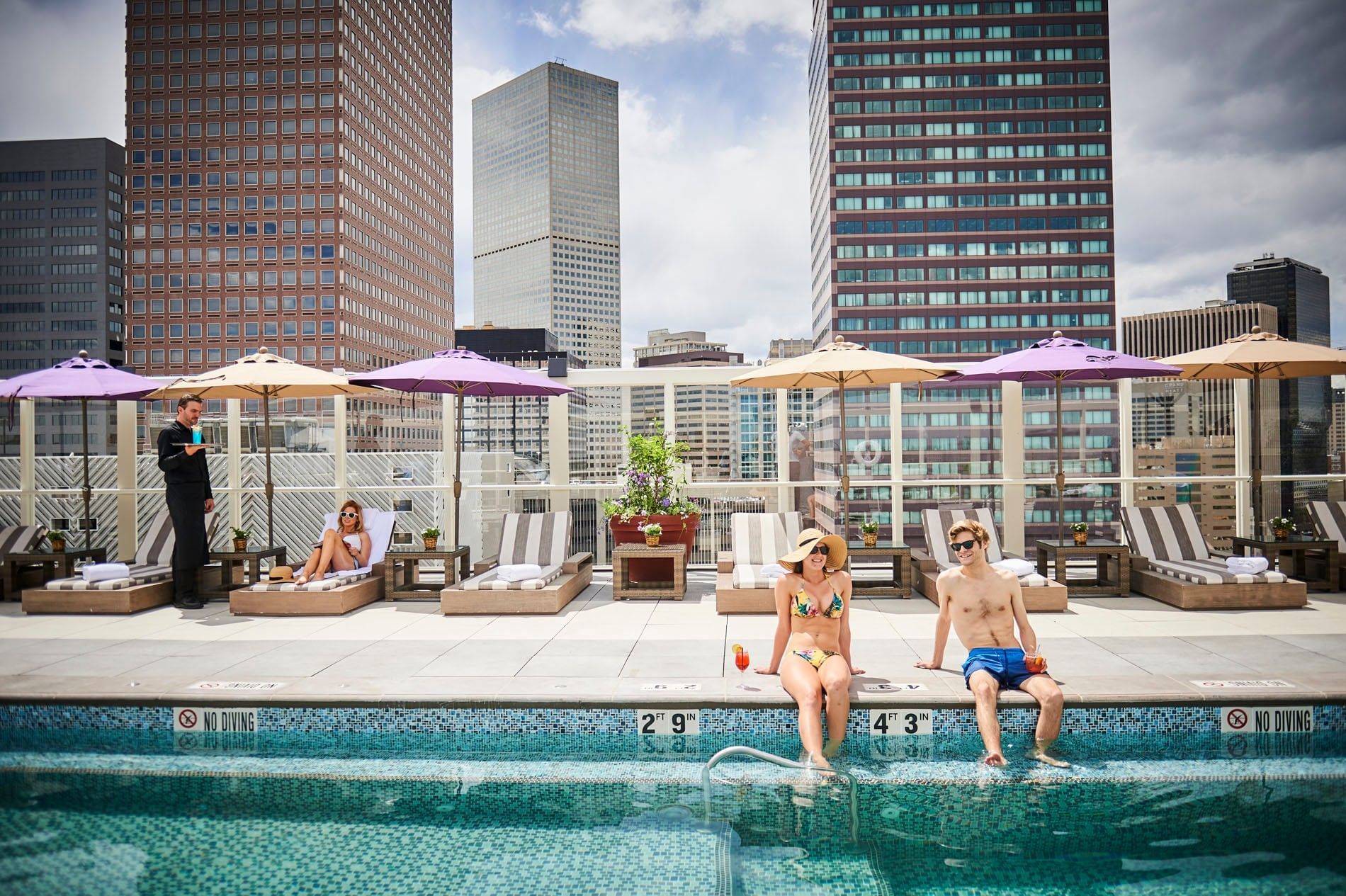 A rooftop pool with lounge chairs and umbrellas, overlooking the city skyline, with people relaxing and enjoying their surroundings.
