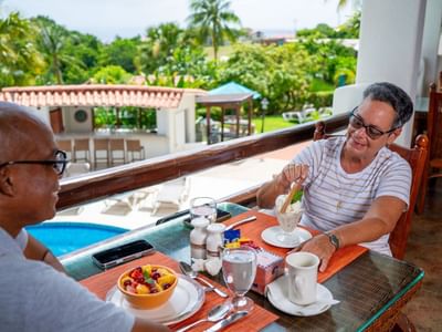 Couple enjoying breakfast on a balcony with a view of a swimming pool and palm trees at Sugar Cane Club Hotel & Spa