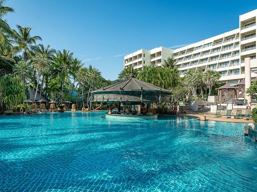 Large outdoor pool with swim-up bar surrounded by palm trees and modern hotel building under clear blue sky at Karon Beach, Phuket