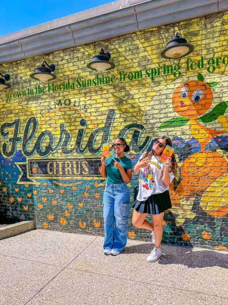 Two girls pose and smile in front of an ornate Famous Florida Citrus mural on a brick wall.