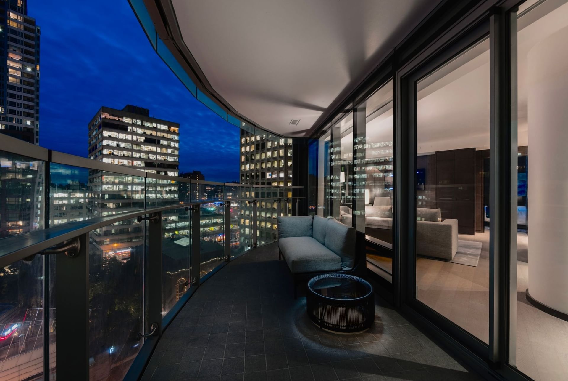Nighttime view of a Paradox Vancouver Hotel balcony with a couch, glass table, and city skyline in the background.