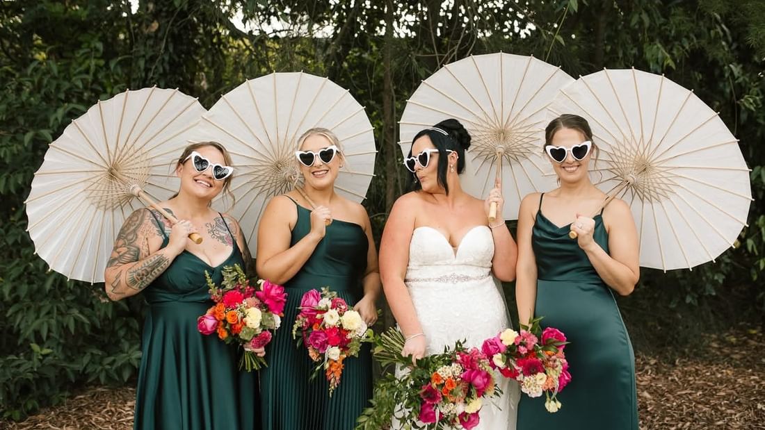 Bride and three bridesmaids in green dresses, holding parasols and bouquets, all wearing heart-shaped sunglasses.