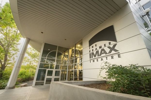 Photograph of an entrance to Kramer IMAX Theatre featuring large glass doors and windows with a white exterior wall displaying bold black signage. Surrounding greenery and a covered walkway with ceiling lights frame the modern architectural design.