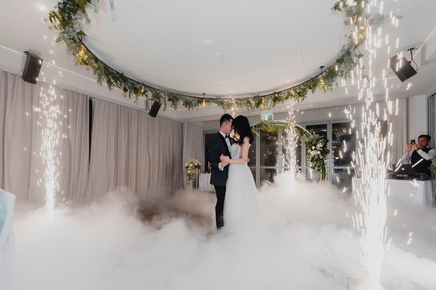 Couple sharing a first dance amid a dreamy, fog-filled room adorned with sparks and greenery at Amora Herencia Riverwalk