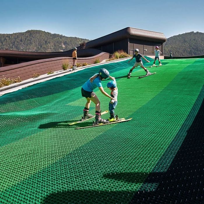 Fünf Skifahrer auf einer grünen Piste mit einem Gebäude und Bergen im Hintergrund.