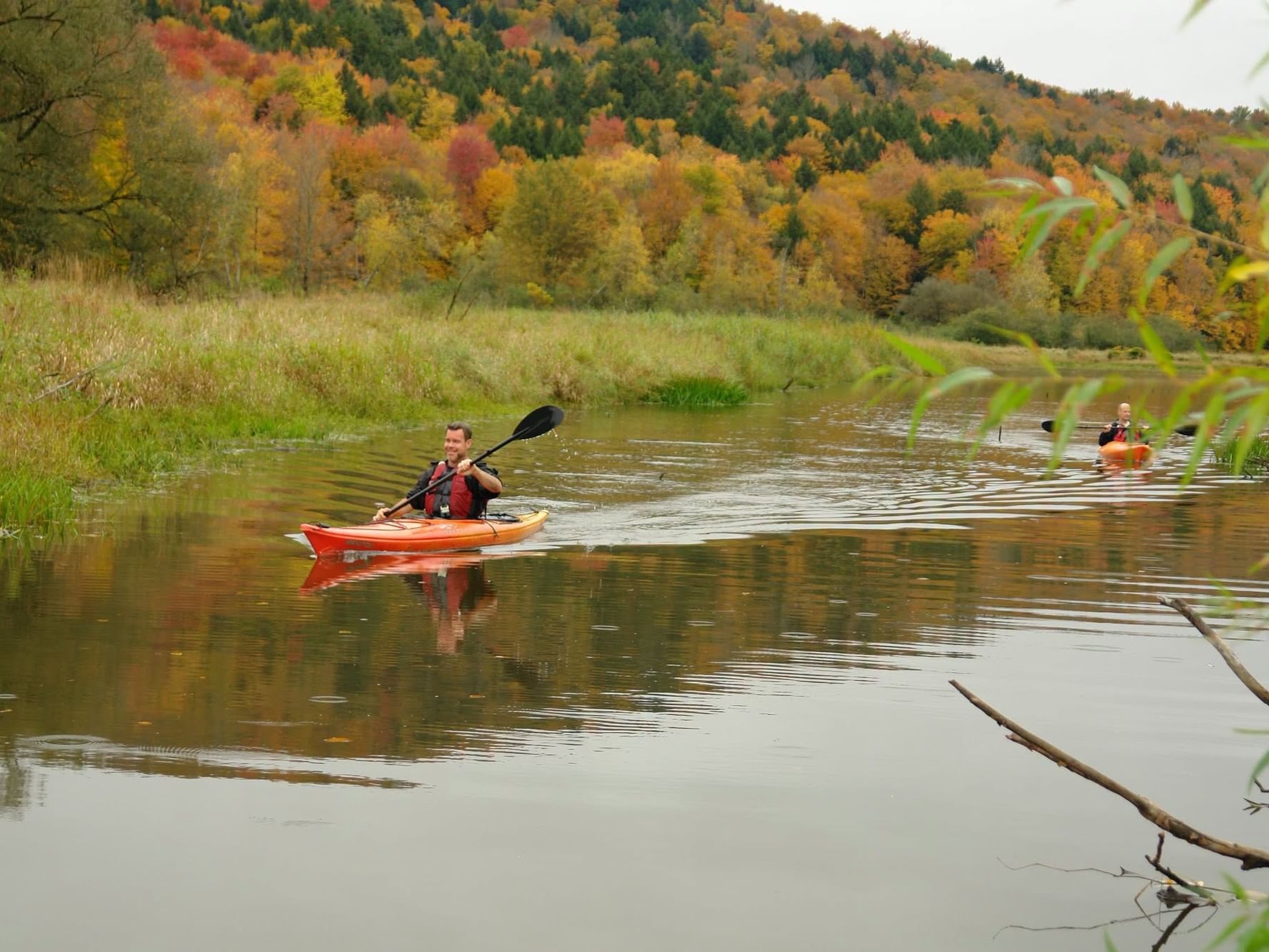 Kayakers paddle along a calm river surrounded by vibrant autumn foliage on hillsides.
