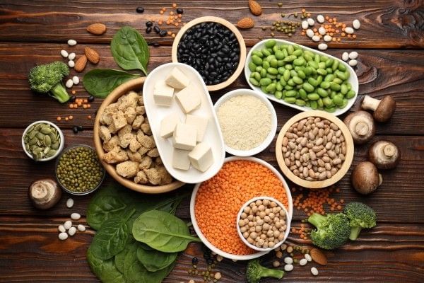 An assortment of plant-based foods against a dark wooden backdrop, including tofu, beans, broccoli, leafy greens, mushrooms, and seeds. 