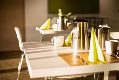 Sunlit breakfast table with a silver coffee carafe, cups, and bright yellow napkins at the Avaneo Hotel Marktredwitz 