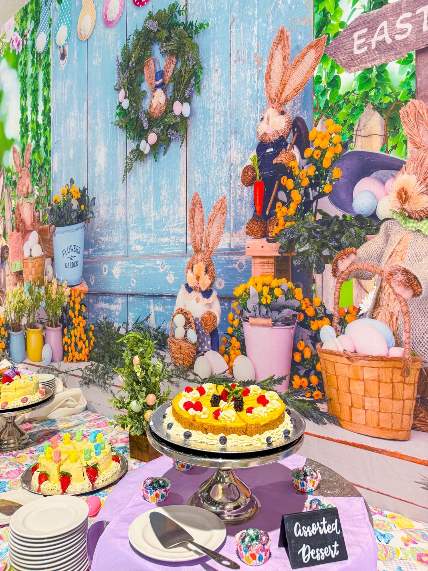 A cake stand with a fruit-decorated cake beside a stack of plates and a sign that reads “Assorted Dessert.” A colorful Easter Bunny backdrop hangs in the background. 