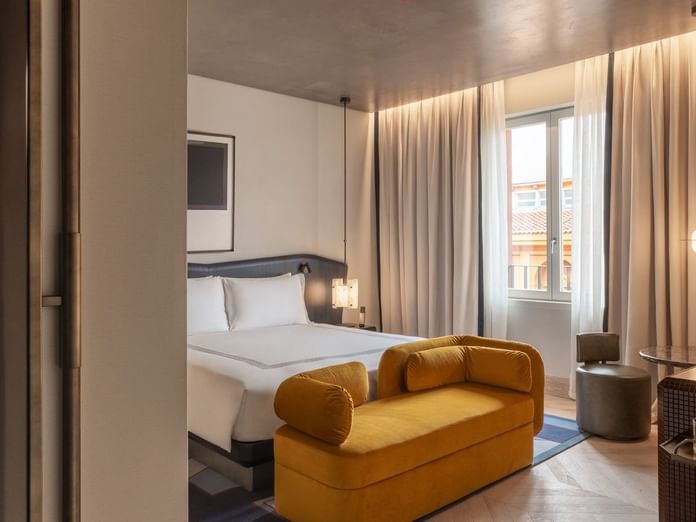Modern guest room with king bed, golden velvet sofa, and balcony doors with city views at The Thompson Rome
