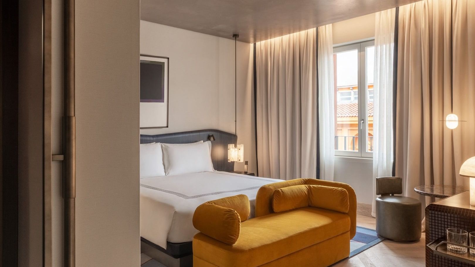 Modern guest room with king bed, golden velvet sofa, and balcony doors with city views at The Thompson Rome