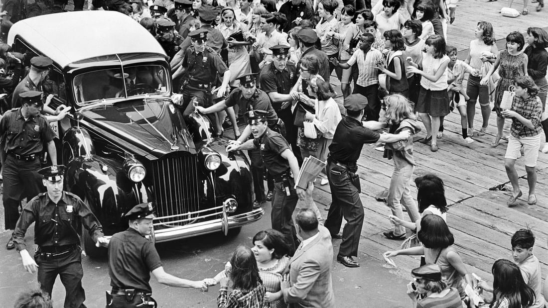 Black and white photograph of a vintage car surrounded by police officers and crowds on a city street during a parade.