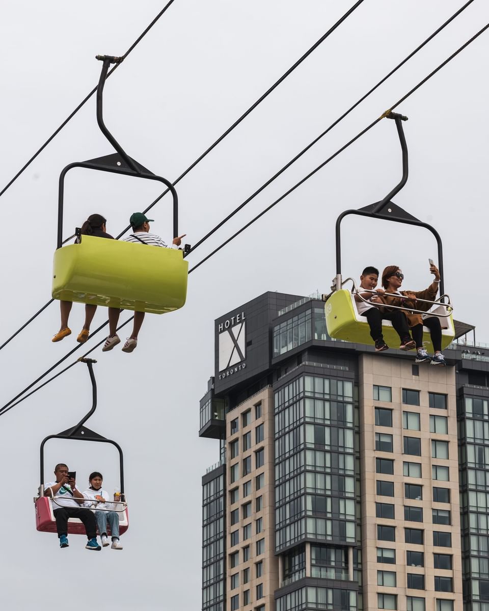 People riding colorful overhead chairlift with modern high-rise building and glass facade behind at Hotel X Toronto