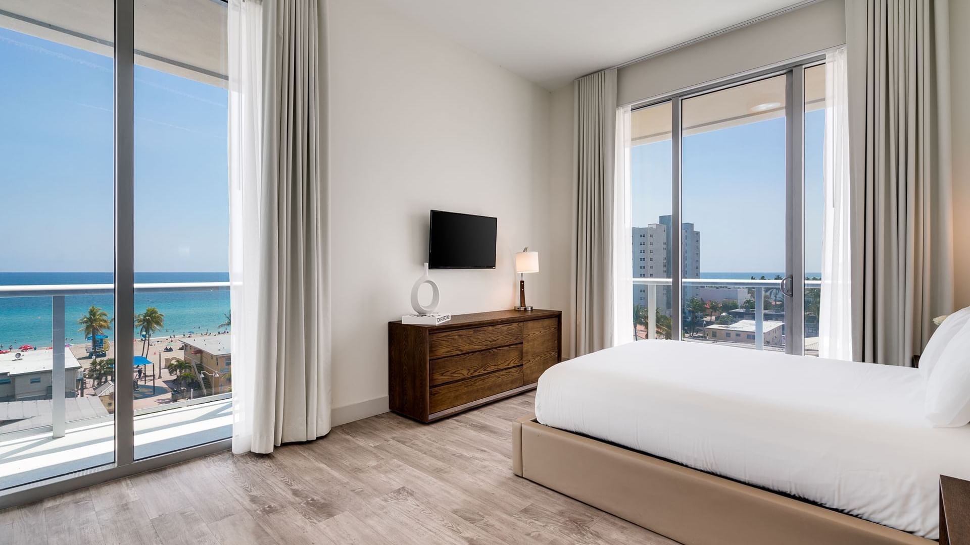 A modern hotel room with a large bed, wooden dresser, and TV, featuring floor-to-ceiling windows with a view of the ocean and balcony.