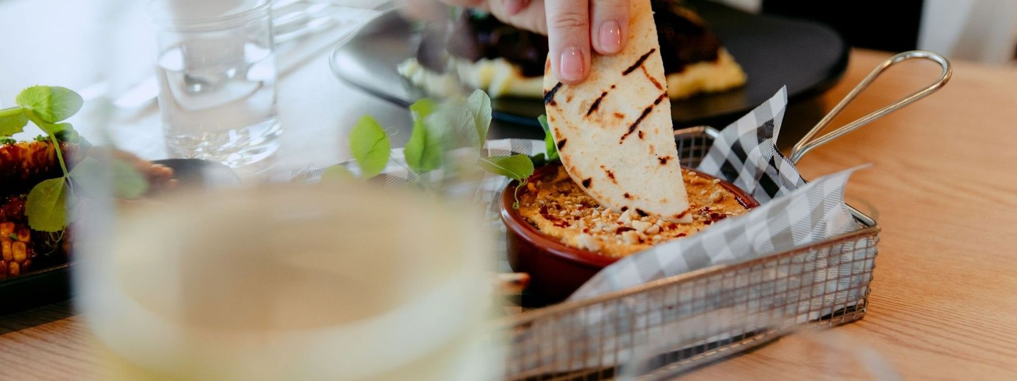 Hand dipping flatbread into hummus bowl with wine glass in foreground promoting Green Neighbour Nights offer.