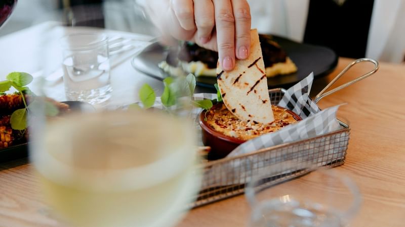 Hand dipping flatbread into hummus bowl with wine glass in foreground promoting Green Neighbour Nights offer.