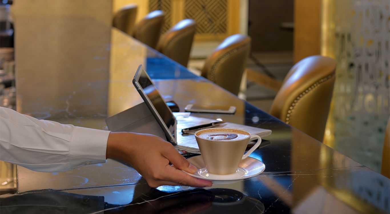 A person reaching for a cup of cappuccino near a laptop at DAMAC Maison Mall Street