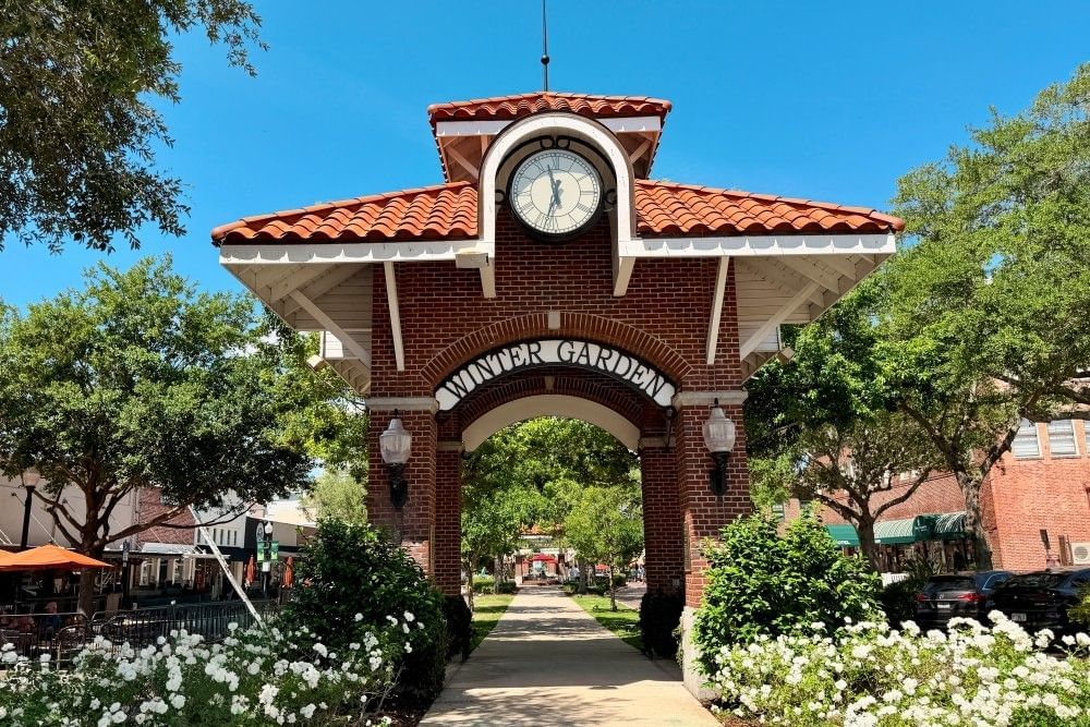 A brick archway reading Winter Garden with a large clock surrounded by mature trees and cobblestone streets lined in shops.