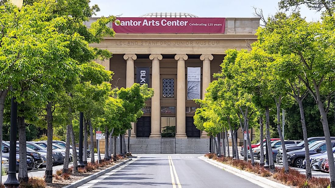 Exterior view of the Grand Cantor Arts Center with a parking lot and rows of green trees near El Prado Hotel