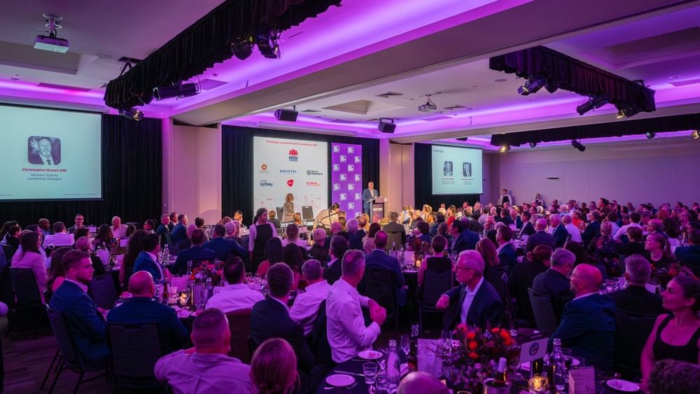 Large banquet hall filled with guests seated at round tables during a formal event with purple ambient lighting and stage.