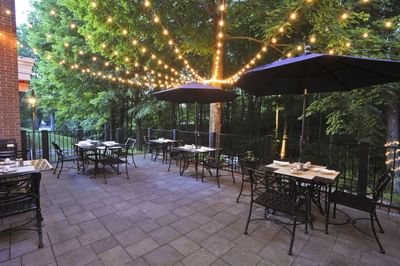 Tables and chairs with umbrellas on a patio facing string lights and trees at Avon Old Farms Hotel