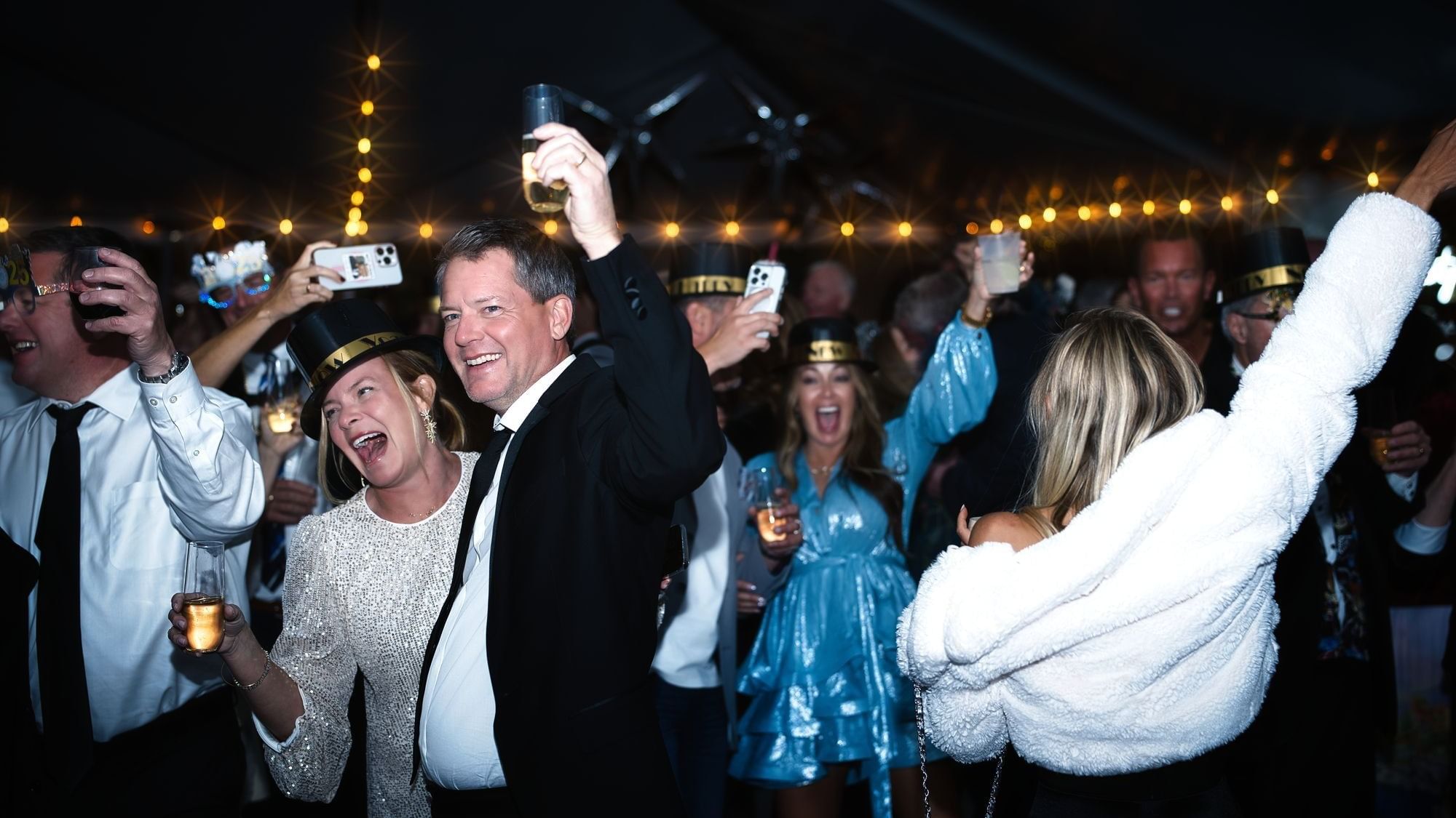 A crowd of people in formal attire, holding drinks, and wearing party hats at a night event.