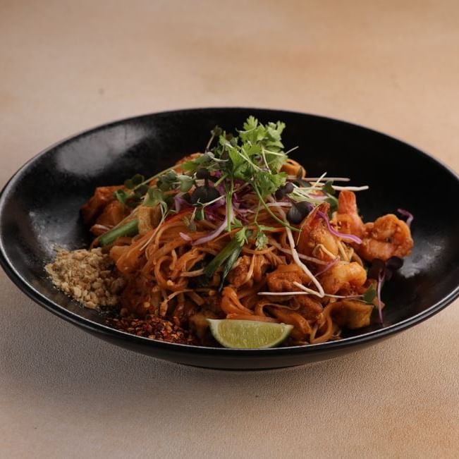 Black bowl with shrimp, noodles, lime wedge, and garnishes on a beige surface.