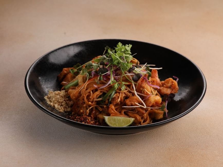 Schwarze Schale mit Garnelen, Nudeln, Limettenspalte und Beilagen auf beigem Untergrund.