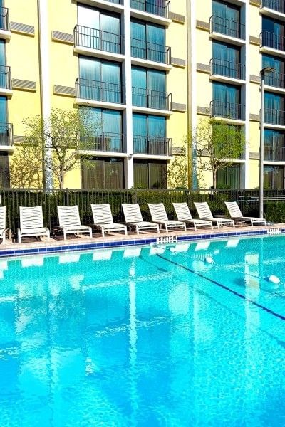 A row of white lounge chairs beside a pool at Rosen Inn, beside a multi-story yellow hotel exterior.
