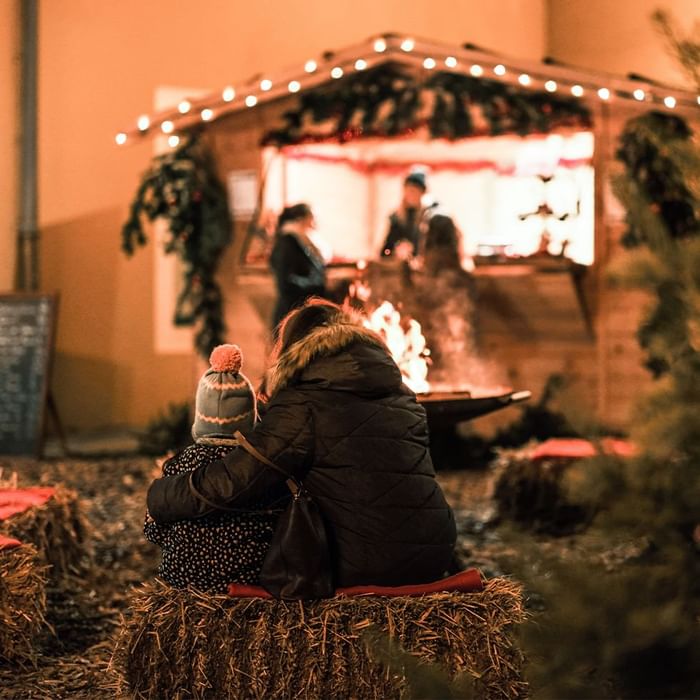 Two people sitting on hay bales by a firepit at a cozy outdoor Christmas market near Falkensteiner Schlosshotel Velden