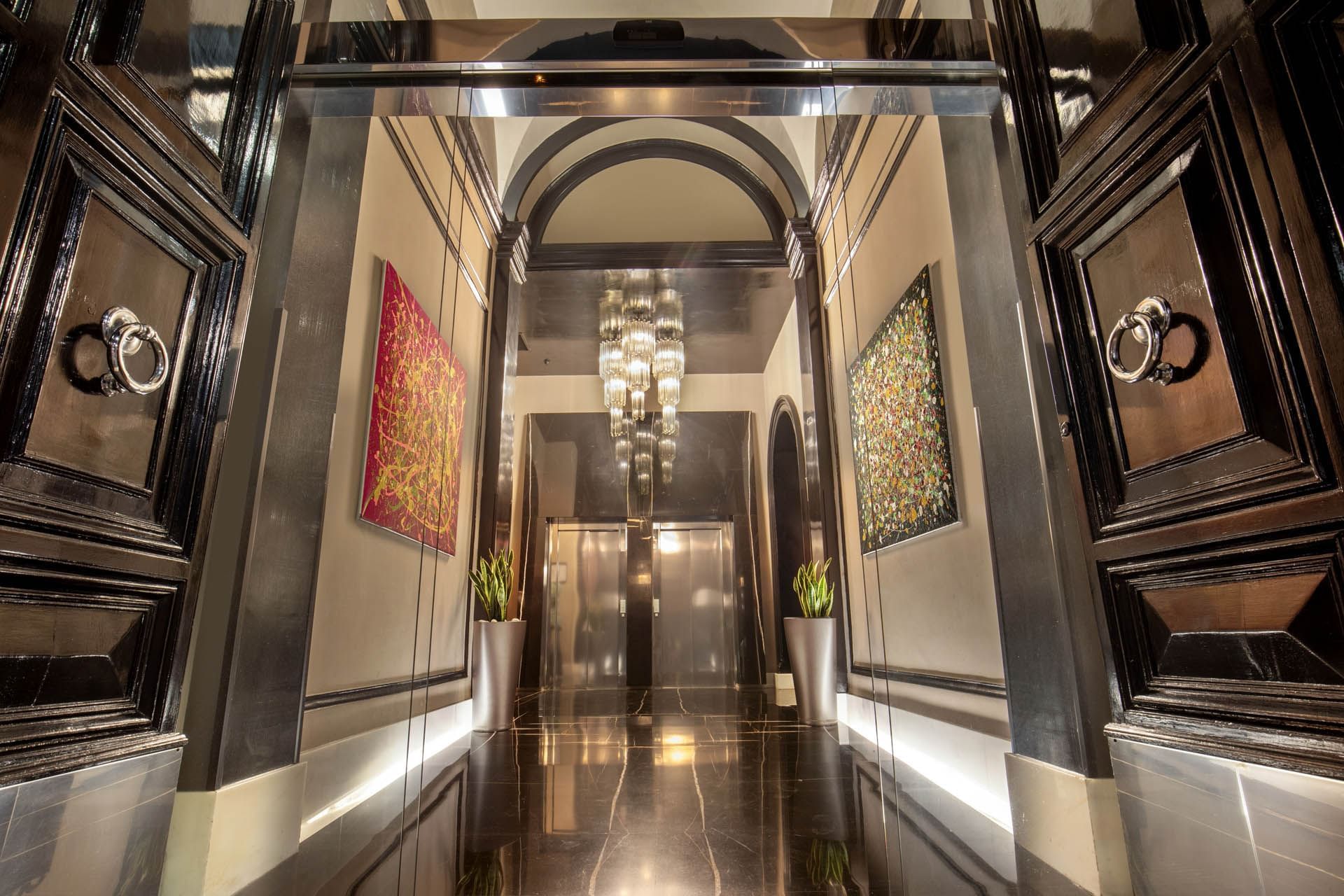Hallway leading to glass elevators, adorned with artwork and a chandelier, framed by glossy black doors at The Glam Hotel