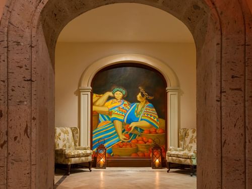 Colorido mural de mujeres con fruta bajo un arco, con dos sillas y faroles en Tomates en Hacienda Del Mar Los Cabos.